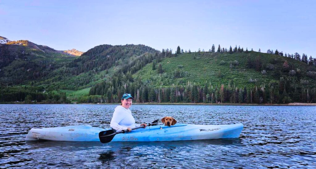 Women sits in kayak with red dog in a blue Utah lake with mountains in background.