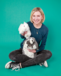 A woman with short blonde hair sits on a teal green backdrop holding a small white bunny in her arms and large white and gray bunny in her lap.