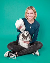 A woman with short blonde hair sits on a teal green backdrop holding a small white bunny in her arms and large white and gray bunny in her lap. 