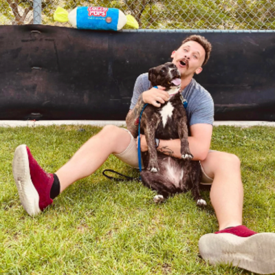 Lolly, a dark brindle dog with a white stripe up her nose, sits in her owner Kev's lap on the ground both have smiling faces.