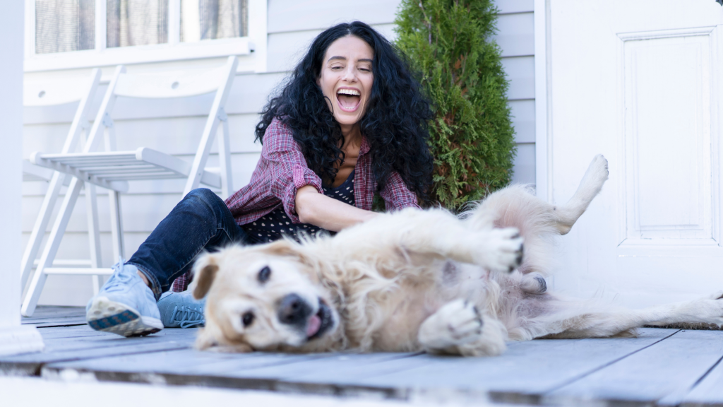 Woman sits on fronch porch smiling while petting a long haired golden colored dog who is laying on his side with his tongue out. 