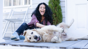 Woman sits on fronch porch smiling while petting a long haired golden colored dog who is laying on his side with his tongue out.