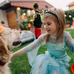 A little girl in a blue princess costume reaches out to pet a large golden colored dog in a yard with halloween decorations.
