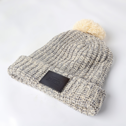 A grey winter hat with cream poof on top and brown leather patch with Humane Society of Utah logo on the base.